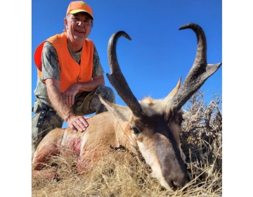 A hunter in an orange vest and camouflage pants kneels beside a pronghorn antelope, captured in a golden field under a clear Wyoming sky during an SNS-guided hunt.